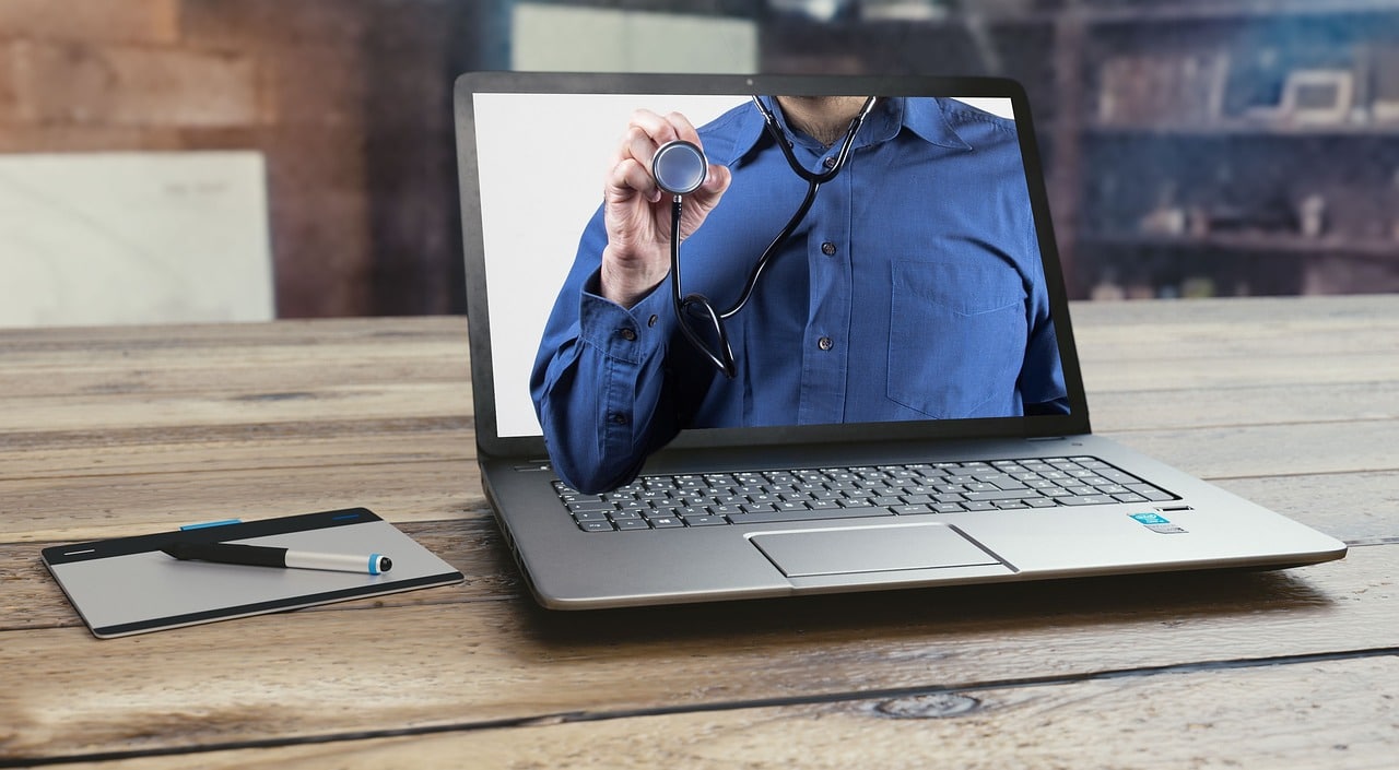 Um laptop sobre uma mesa de madeira exibe a imagem de uma pessoa de camisa azul segurando um estetoscópio ilustrando a telessaúde