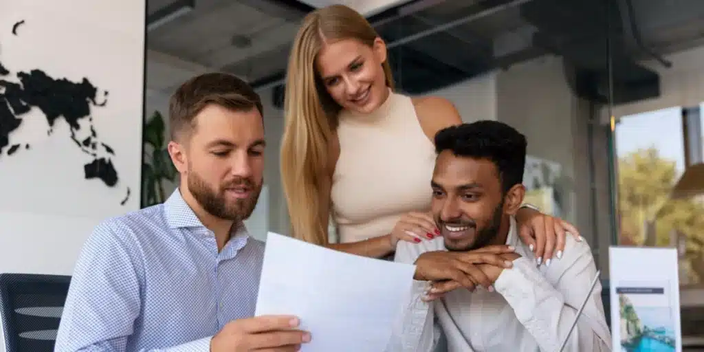 Três pessoas estão reunidas em um escritório. Um homem barbudo segura um documento rotulado Benefícios Corporativos, enquanto uma mulher e outro homem olham e sorriem. Eles parecem estar discutindo a papelada. Um mapa-múndi adorna a parede atrás deles, adicionando um toque global à conversa.