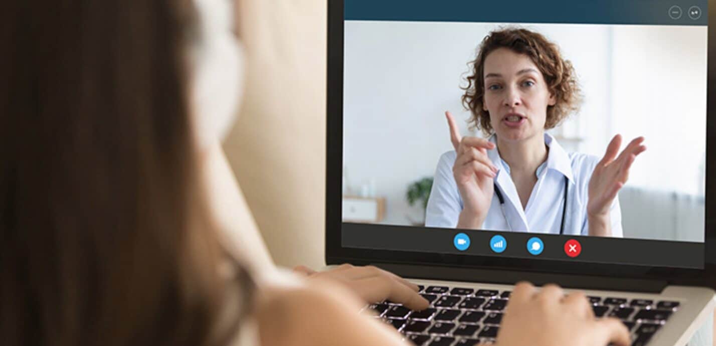 Uma pessoa em videochamada exibida na tela de um laptop, com outra pessoa assistindo de trás, ilustrando a telemedicina para empresas