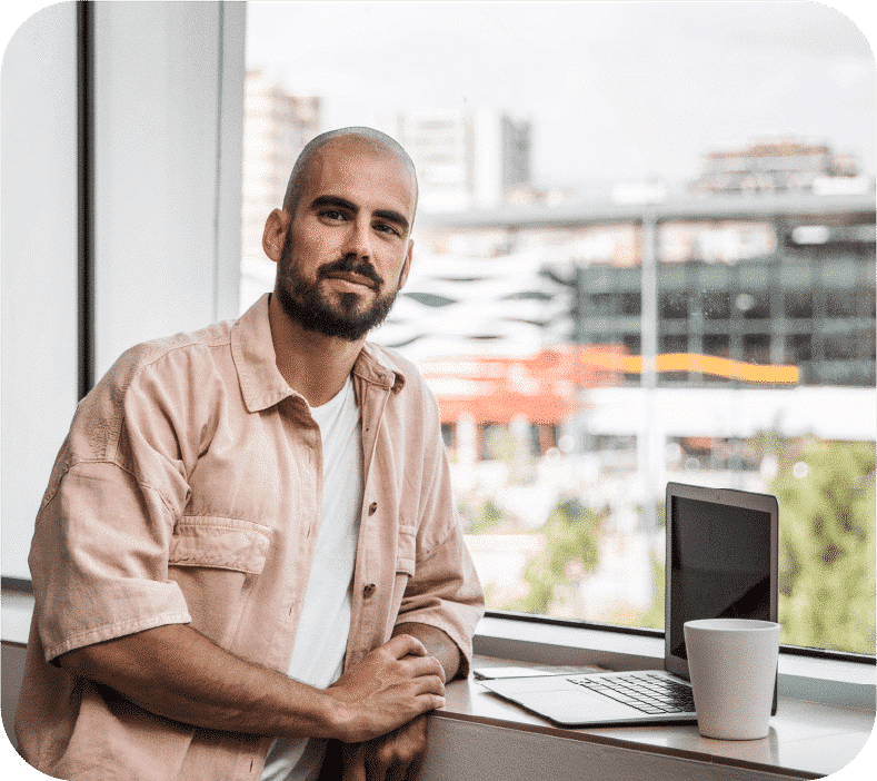 Um homem olhando para a câmera na frente dele tem um laptop e uma caneca branca e ao fundo uma paisagem urbana