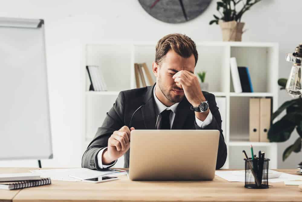 Um homem de terno está sentado em uma mesa com um laptop, parecendo estressado representando a importância da prevenção de doenças no ambiente de trabalho