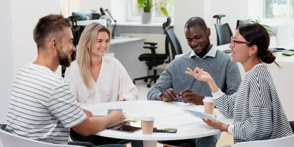 Quatro pessoas sentam-se ao redor de uma mesa em um escritório iluminado, envolvidas em comunicação assertiva. Elas seguram papéis e dispositivos eletrônicos enquanto tomam café. A sala tem móveis modernos e janelas grandes, criando um ambiente ideal para discussões eficazes no local de trabalho.