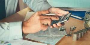 Uma pessoa calcula meticulosamente com uma caneta na mão, cercada por documentos financeiros e pilhas de moedas em uma mesa de madeira, incorporando eficiência tributária com precisão. Perto, um jarro de vidro transbordando de moedas reflete sua dedicação à gestão inteligente de recursos.