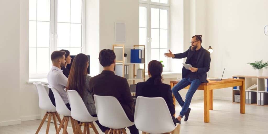 Homem sentando em uma mesa dando uma apresentação para um grupo de cinco pessoas, representando programas de bem-estar para empresas