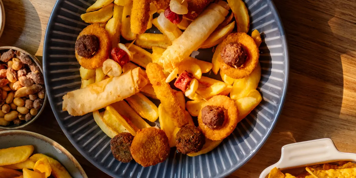 Prato com porção de batatas fritas e acompanhamentos, incluindo rodelas empanadas e pedaços crocantes sobre uma mesa de madeira, com tigelas laterais de grãos e molho.