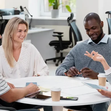 Quatro pessoas estão reunidas em volta de uma mesa em um escritório, envolvidas em uma discussão que reflete comunicação assertiva no trabalho. Três delas estão sorrindo, contribuindo para a atmosfera positiva. Papéis e xícaras de café espalhados pela mesa, enquanto uma luz brilhante ilumina cadeiras vazias e plantas ao fundo.