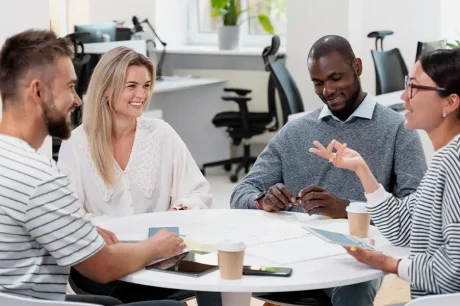 Quatro pessoas estão reunidas em volta de uma mesa em um escritório, envolvidas em uma discussão que reflete comunicação assertiva no trabalho. Três delas estão sorrindo, contribuindo para a atmosfera positiva. Papéis e xícaras de café espalhados pela mesa, enquanto uma luz brilhante ilumina cadeiras vazias e plantas ao fundo.