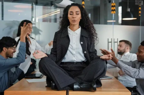 Colaboradores em uma mesa no ambiente de trabalho tendo discussões, com estresse e sobrecarga, mas uma colaboradora está sentada em cima da mesa em posição de lotus pela yoga, fazendo uma meditação e tentando se acalmar. Impacto da NR-01 nas empresas são significativos e de extrema importância para produtividade, saúde e bem-estar do time