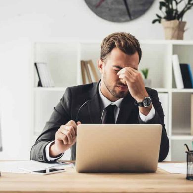 Um homem de terno está sentado em uma mesa com um laptop, parecendo estressado representando a importância da prevenção de doenças no ambiente de trabalho