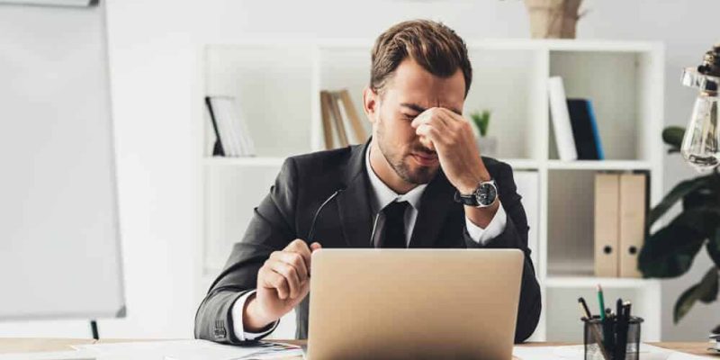 Um homem de terno está sentado em uma mesa com um laptop, parecendo estressado representando a importância da prevenção de doenças no ambiente de trabalho