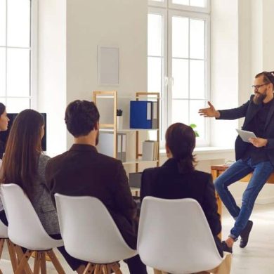 Homem sentando em uma mesa dando uma apresentação para um grupo de cinco pessoas, representando programas de bem-estar para empresas