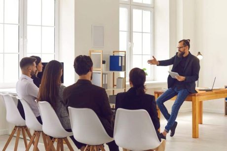 Homem sentando em uma mesa dando uma apresentação para um grupo de cinco pessoas, representando programas de bem-estar para empresas