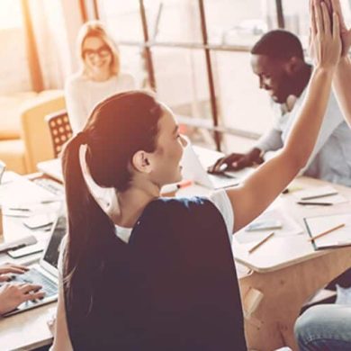 Em um ambiente de escritório, um grupo colabora em volta de uma mesa com laptops, papéis e outras pessoas. Ilustra a saúde emocional no ambiente de corporativo