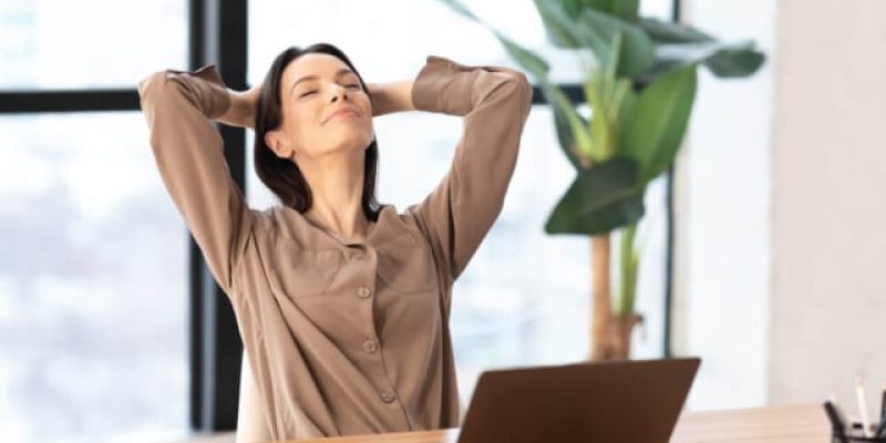 Uma mulher de blusa bege se recosta na cadeira com as mãos atrás da cabeça, personificando a essência da saúde mental no trabalho.