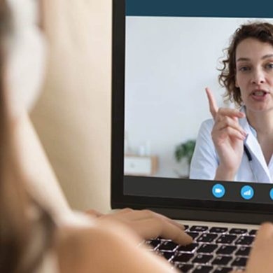 Uma pessoa em videochamada exibida na tela de um laptop, com outra pessoa assistindo de trás, ilustrando a telemedicina para empresas