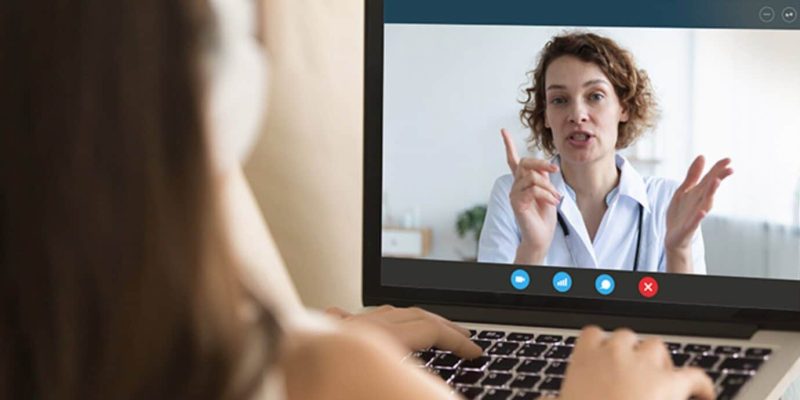 Uma pessoa em videochamada exibida na tela de um laptop, com outra pessoa assistindo de trás, ilustrando a telemedicina para empresas