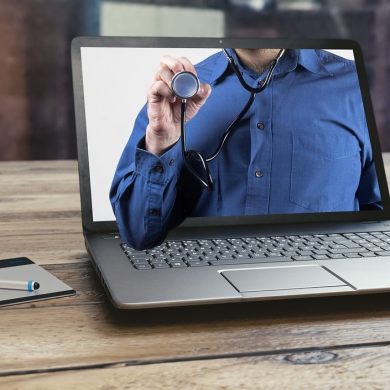 Um laptop sobre uma mesa de madeira exibe a imagem de uma pessoa de camisa azul segurando um estetoscópio ilustrando a telessaúde no Brasil