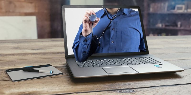 Um laptop sobre uma mesa de madeira exibe a imagem de uma pessoa de camisa azul segurando um estetoscópio ilustrando a telessaúde